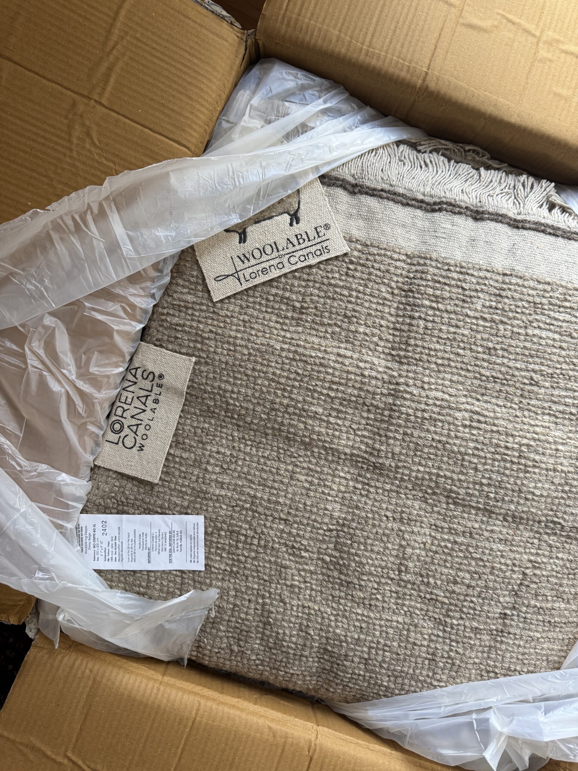 A beige Lorena Canals wool rug partially wrapped in plastic is shown inside an open cardboard box, with product tags and care labels visible.