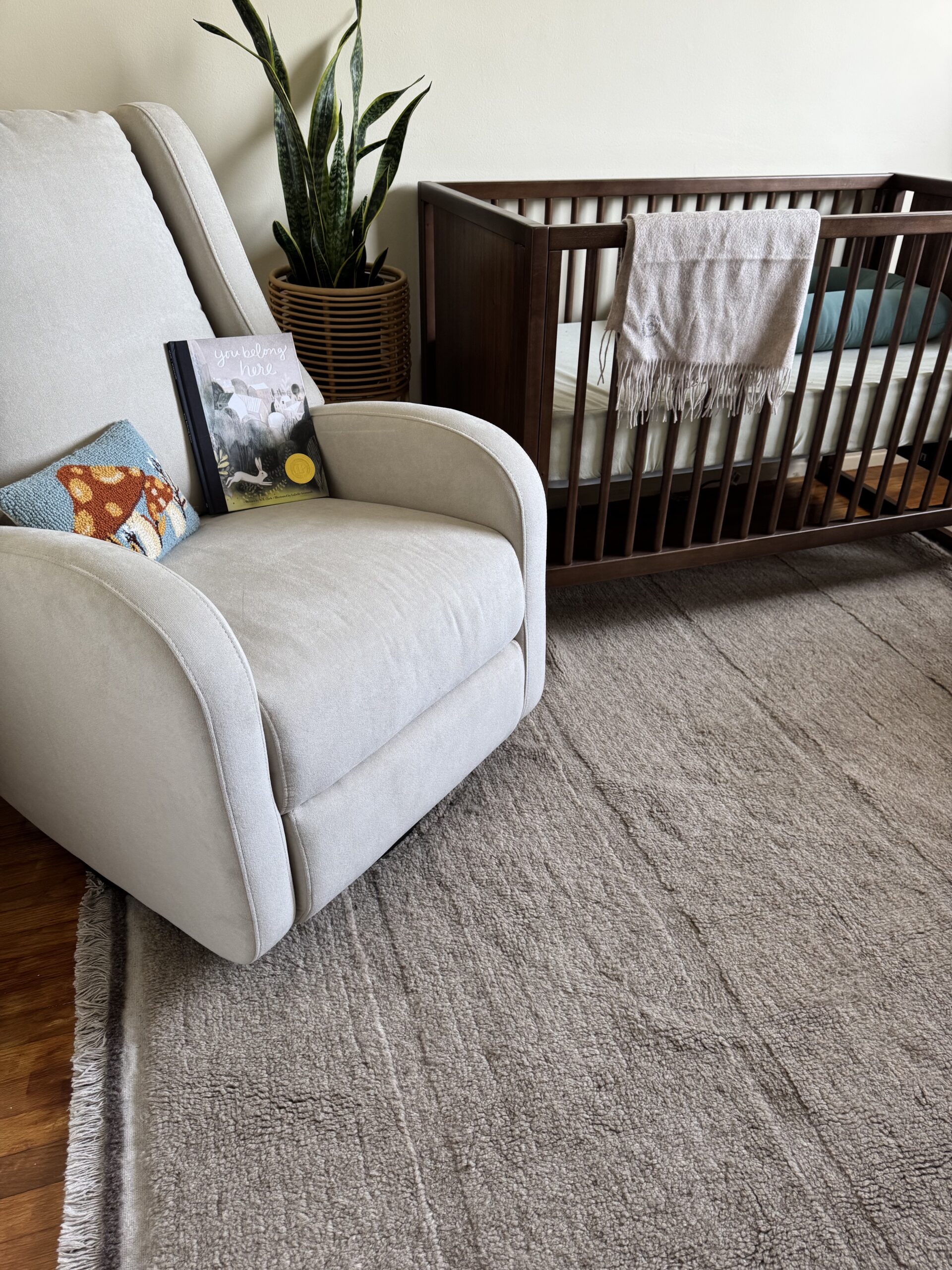 A beige recliner with a pillow and children's book beside a wooden crib with a light blanket draped over the railing, and a potted plant in the corner, all on a neutral rug.