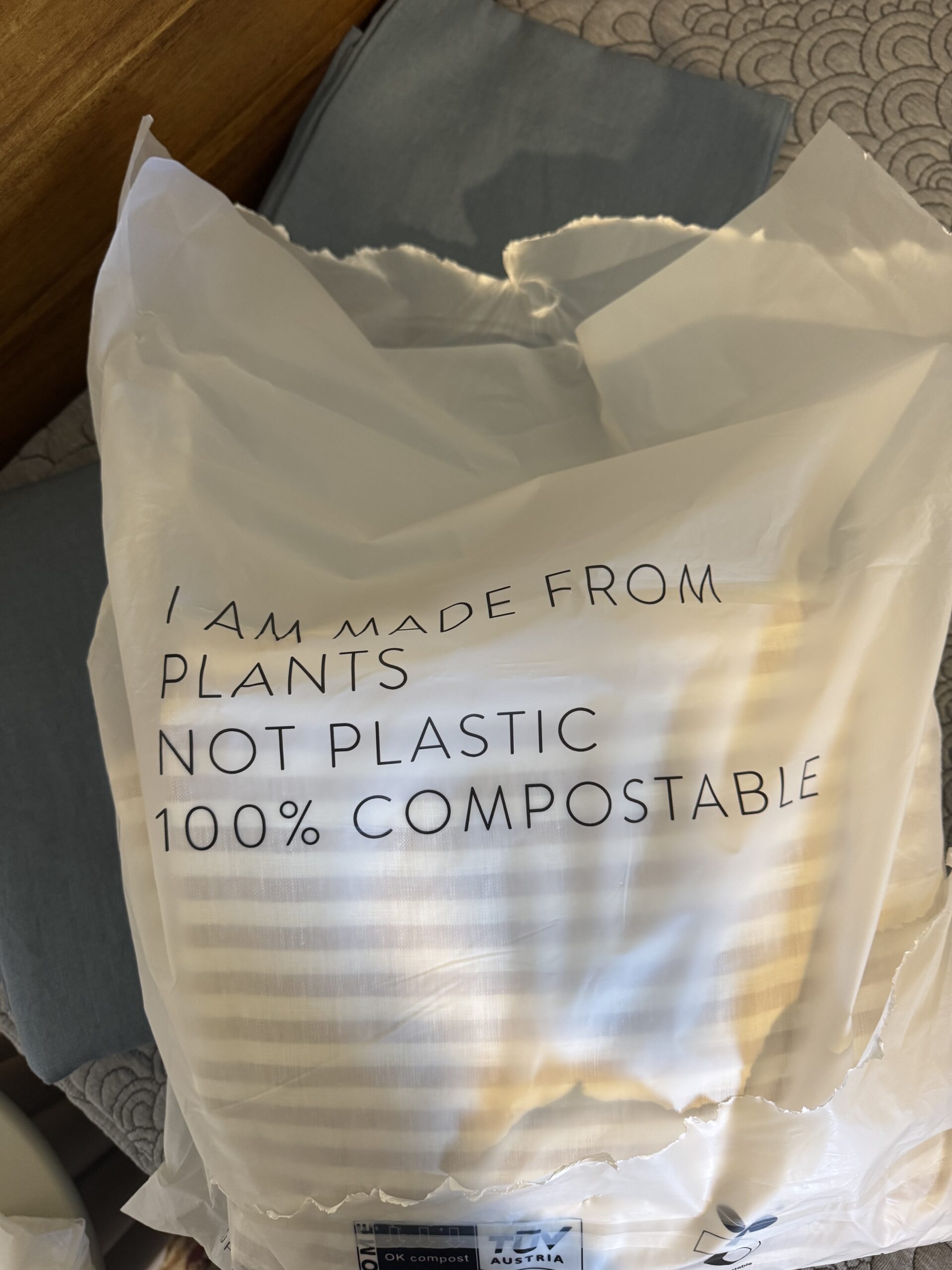 A white bag with black text reads, "I am made from plants not plastic 100% compostable," placed on a bed with pillows.