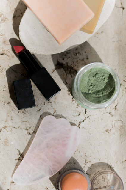 A flat lay of beauty products including a red lipstick, green powder in a jar, a pink gua sha stone, a round cream blush, and two soap bars on a beige surface.