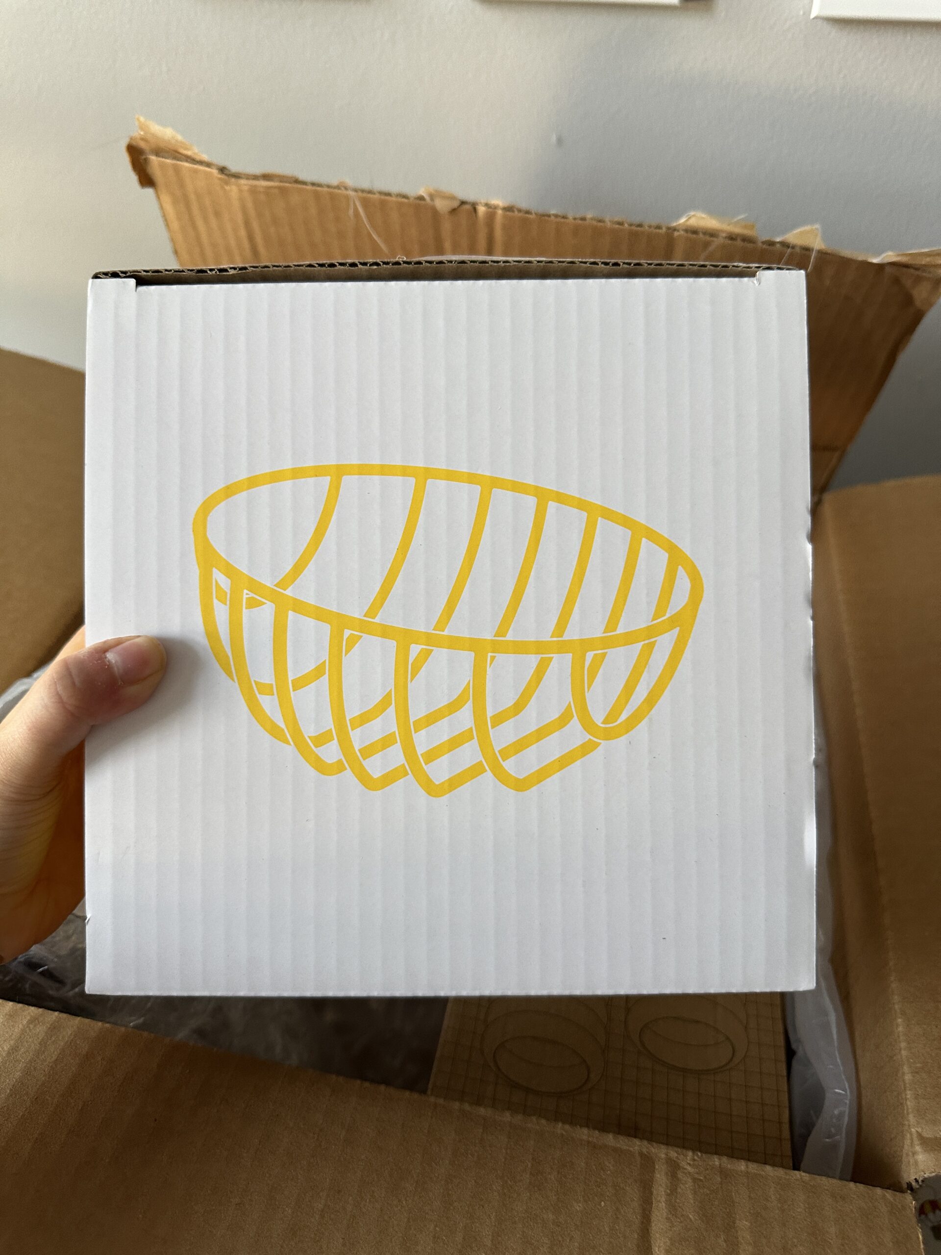 A hand holds a white cardboard box with a yellow line drawing of a bowl on its front, positioned above an open brown shipping box.