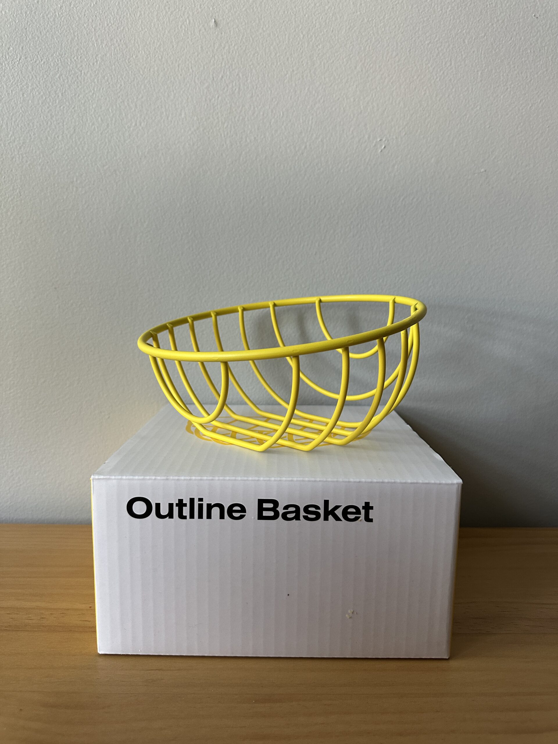 A yellow wire basket sits on top of a white box labeled "Outline Basket" on a wooden surface against a plain wall.