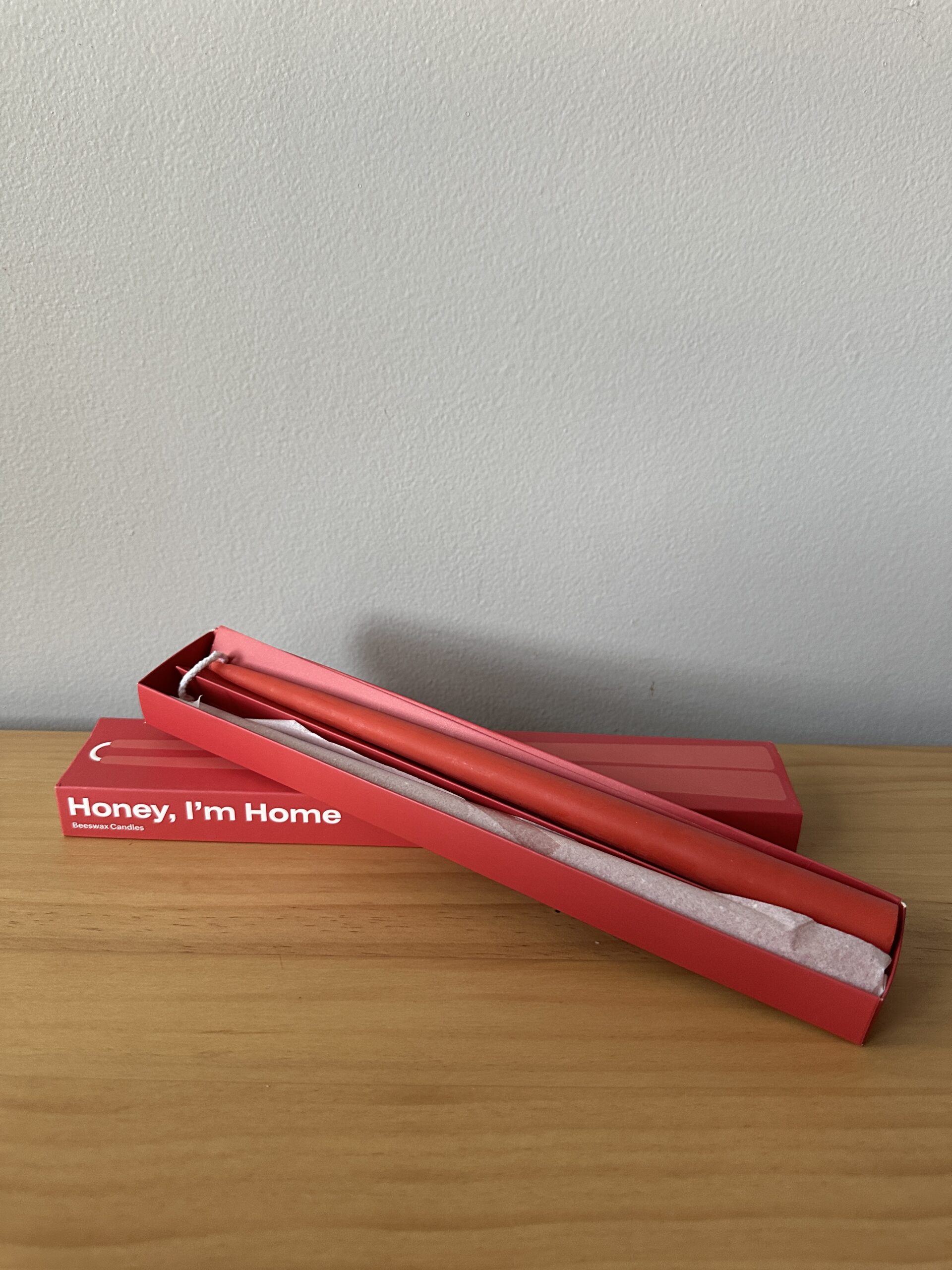 A red box labeled "Honey, I'm Home" lies open on a wooden surface, revealing a red candle inside.
