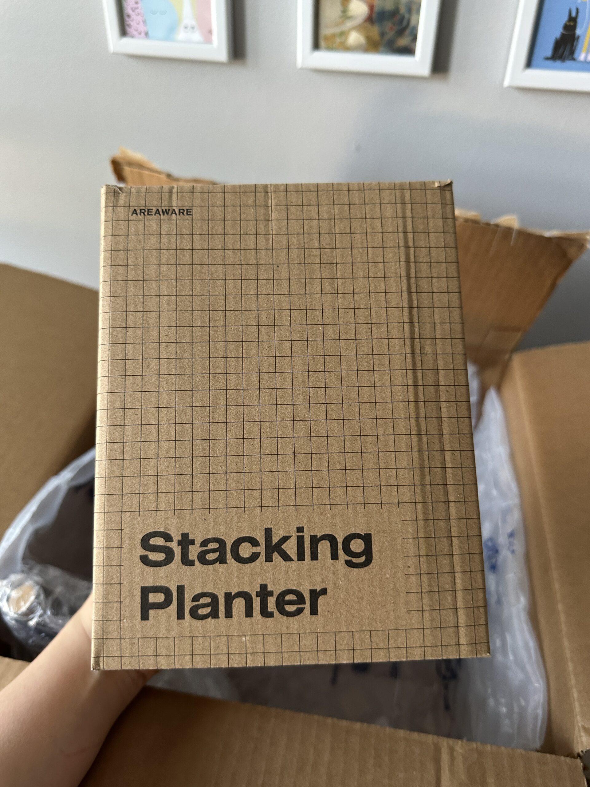 A hand holding a cardboard box labeled "Stacking Planter" in front of an open shipping box, with framed pictures on the wall in the background.
