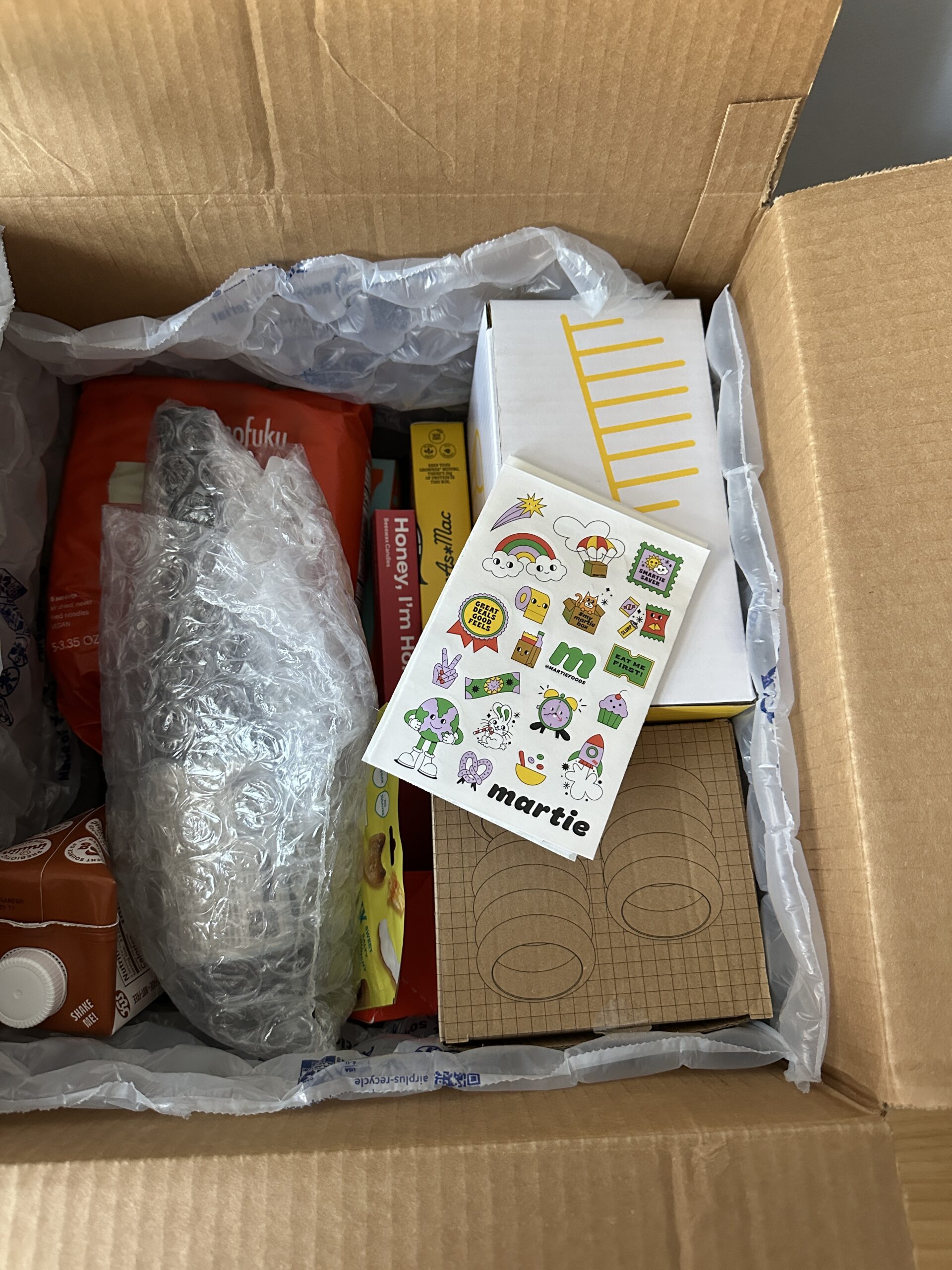 An open cardboard box containing assorted packaged items, a bubble-wrapped object, and a sheet of colorful stickers labeled "martie.
