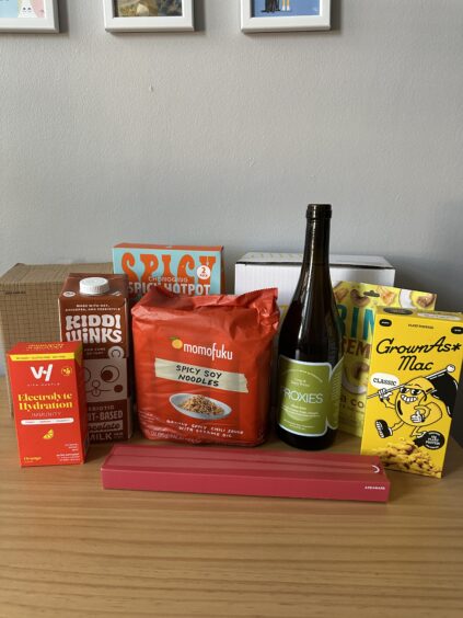 Assorted food and drink items, including hydration mix, chocolate milk, spicy soy noodles, a wine bottle, mac and cheese, and a red rectangular box, displayed on a wooden surface.