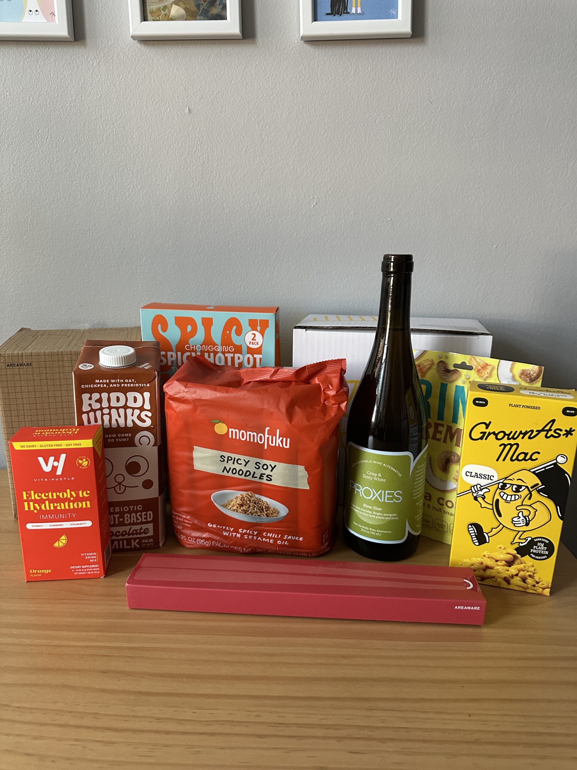 Assorted food and drink items, including hydration mix, chocolate milk, spicy soy noodles, a wine bottle, mac and cheese, and a red rectangular box, displayed on a wooden surface.