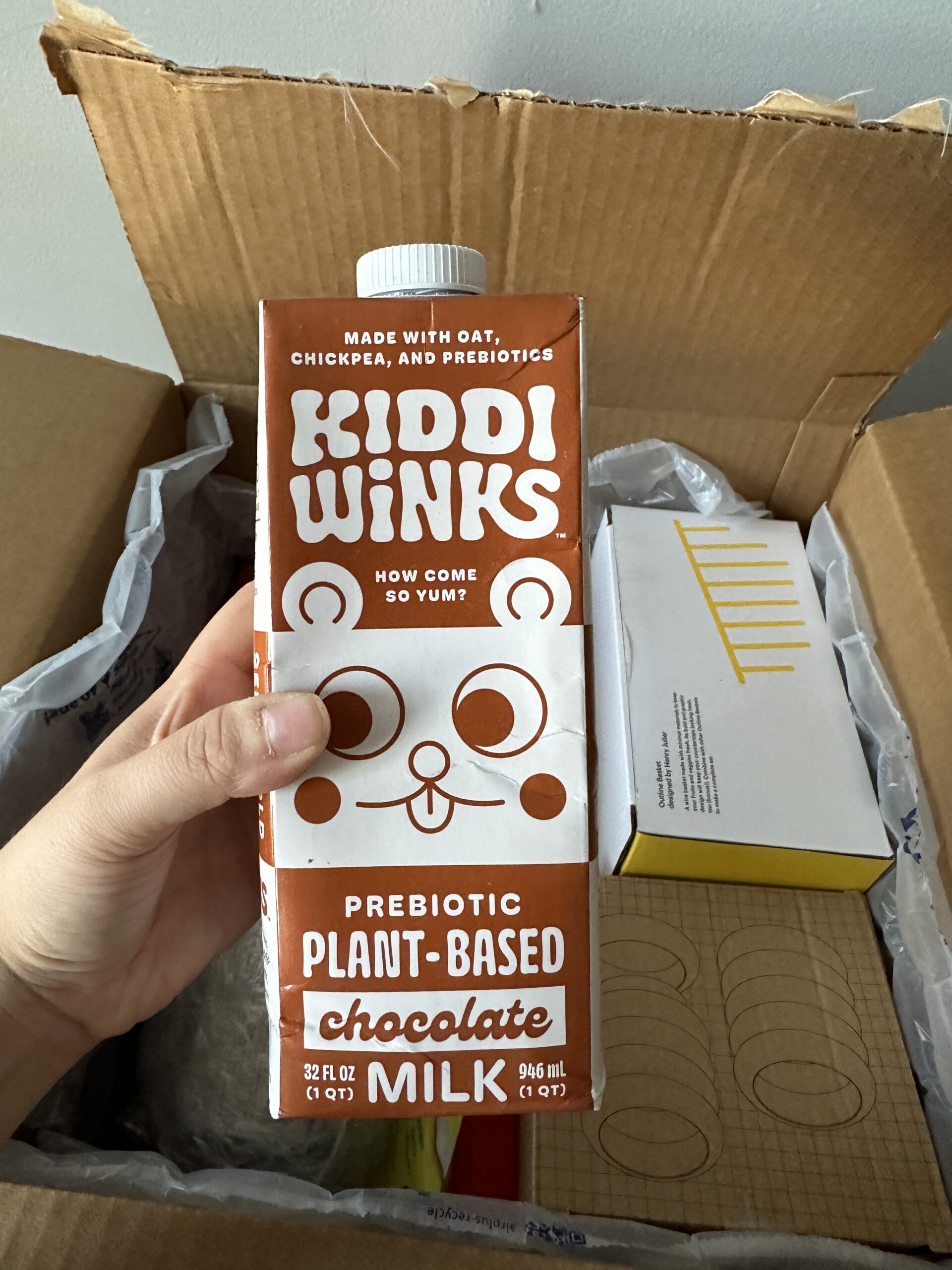A hand holds a carton of Kiddi Winks prebiotic plant-based chocolate milk over an open cardboard box.