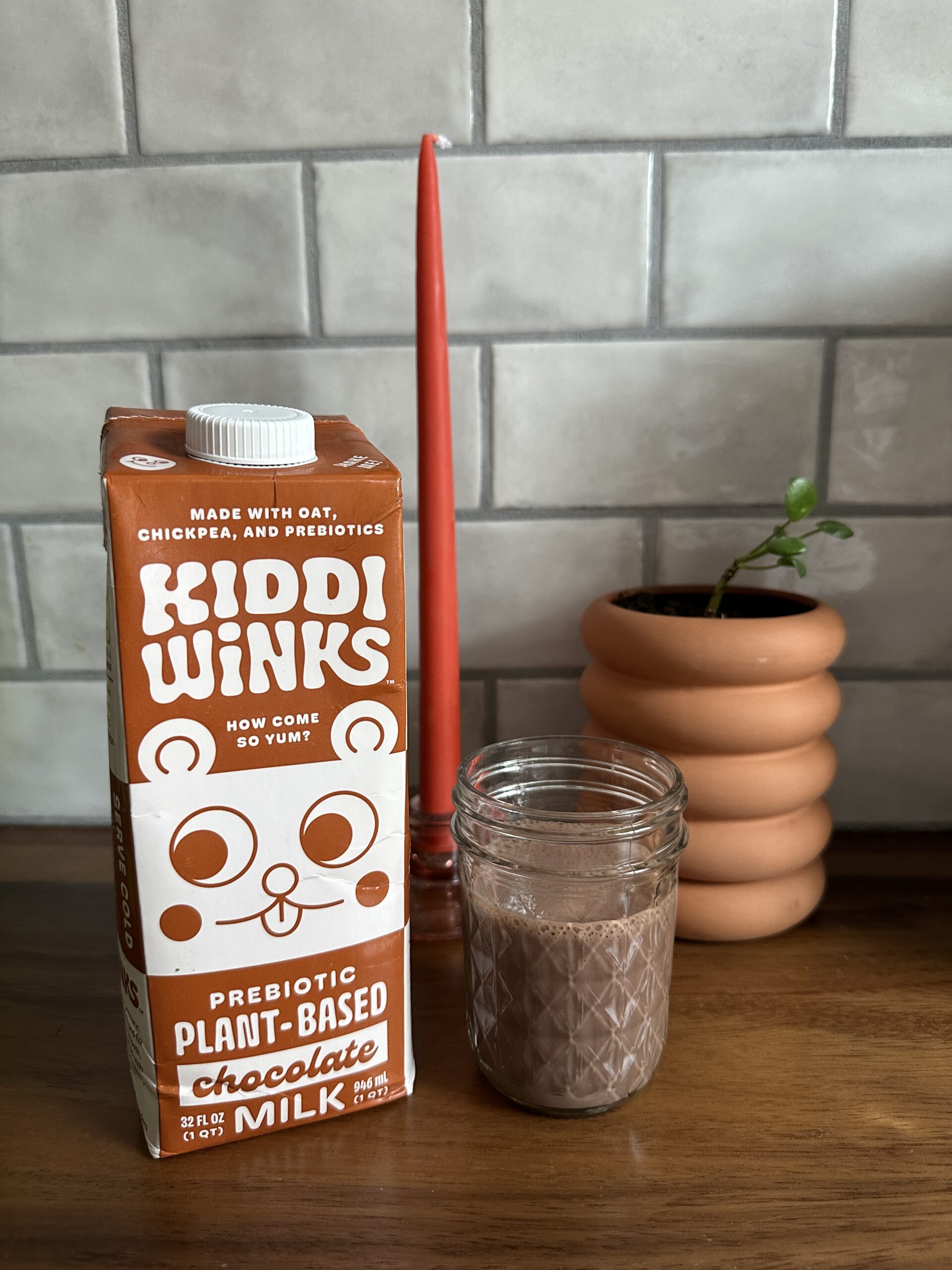 A carton of Kiddi Winks prebiotic plant-based chocolate milk sits next to a glass of chocolate milk, a red candle, and a small potted plant on a wooden surface.