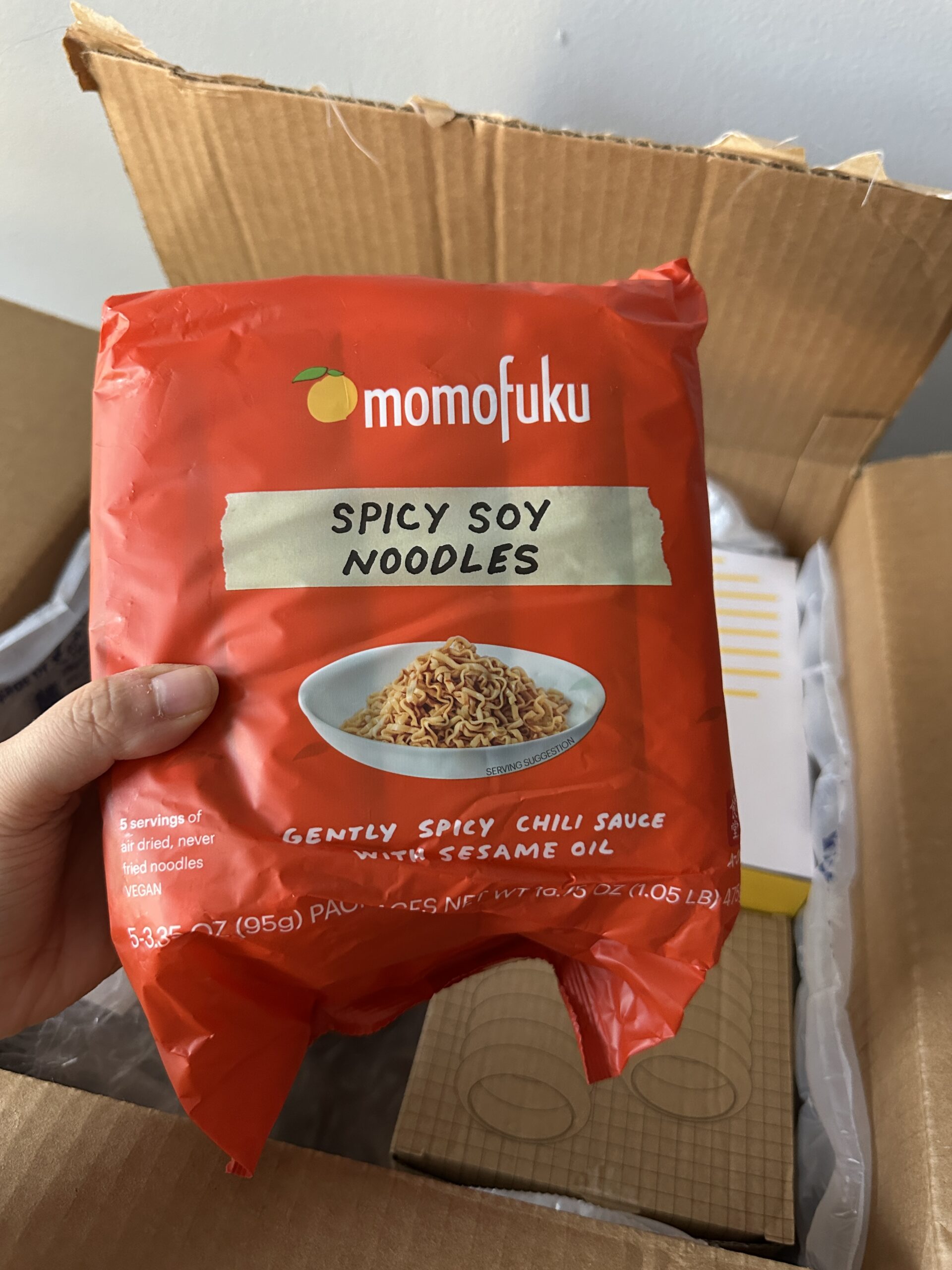 A hand holds a pack of Momofuku Spicy Soy Noodles in front of an open cardboard box.