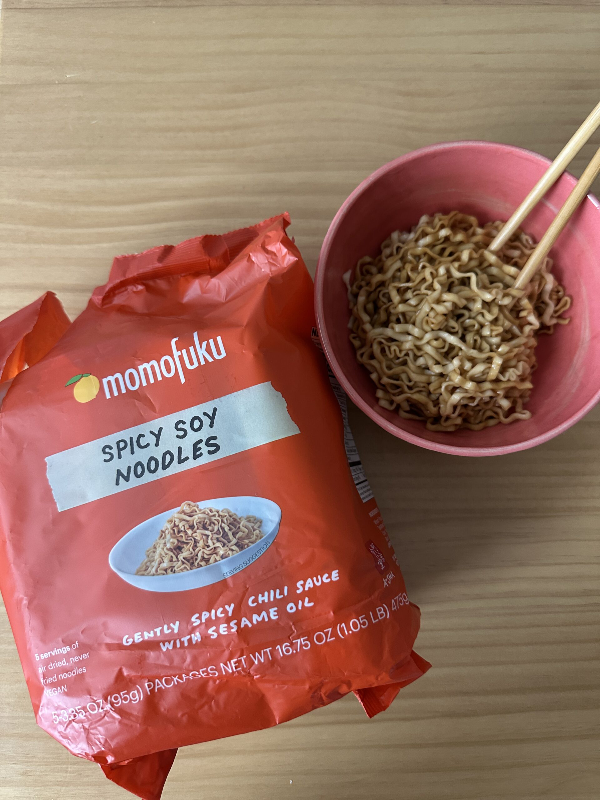 A package of Momofuku Spicy Soy Noodles next to a pink bowl filled with cooked noodles and a pair of wooden chopsticks on a wooden surface.