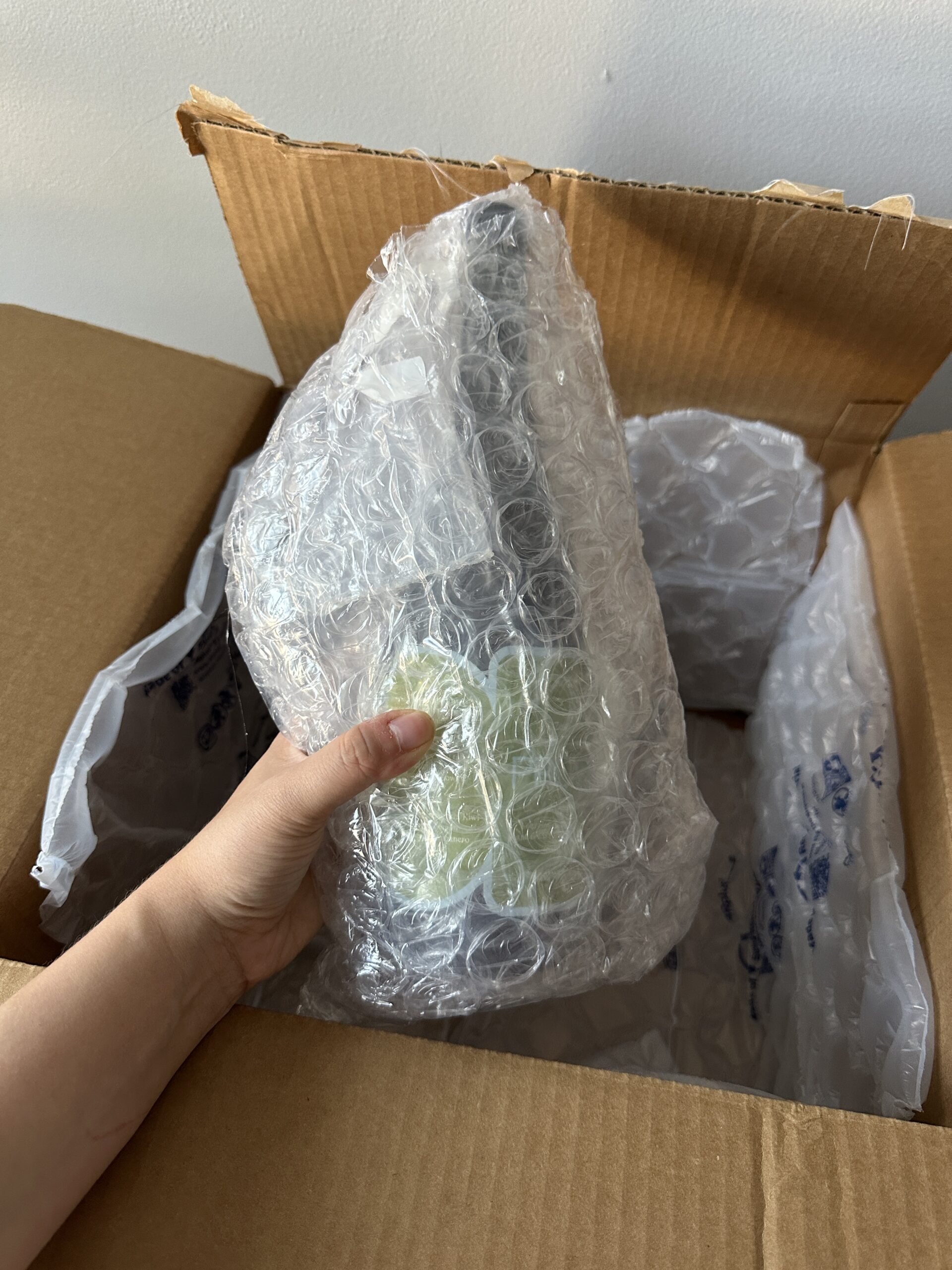 A hand holds a bubble-wrapped item inside an open cardboard shipping box with air-filled packing material.