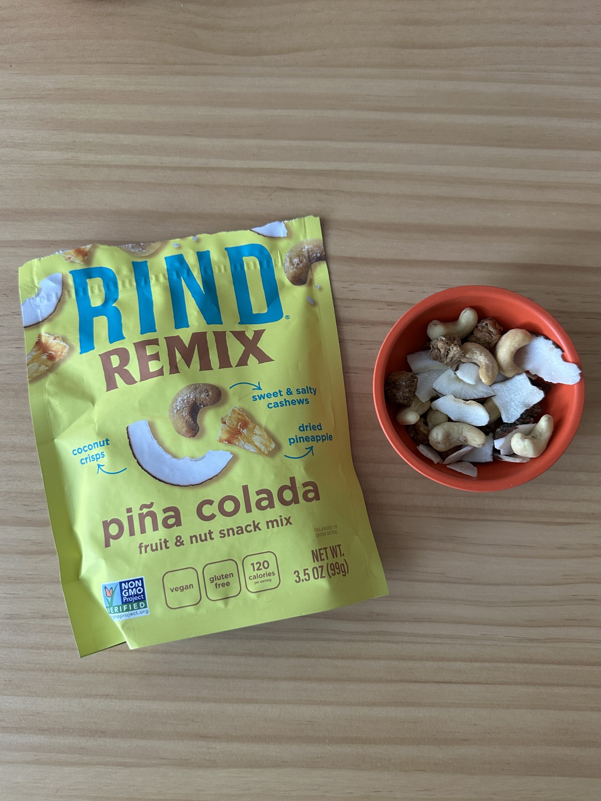 A package of RIND Remix Piña Colada fruit and nut snack mix is next to a small orange bowl containing the snack mix, on a wooden surface.