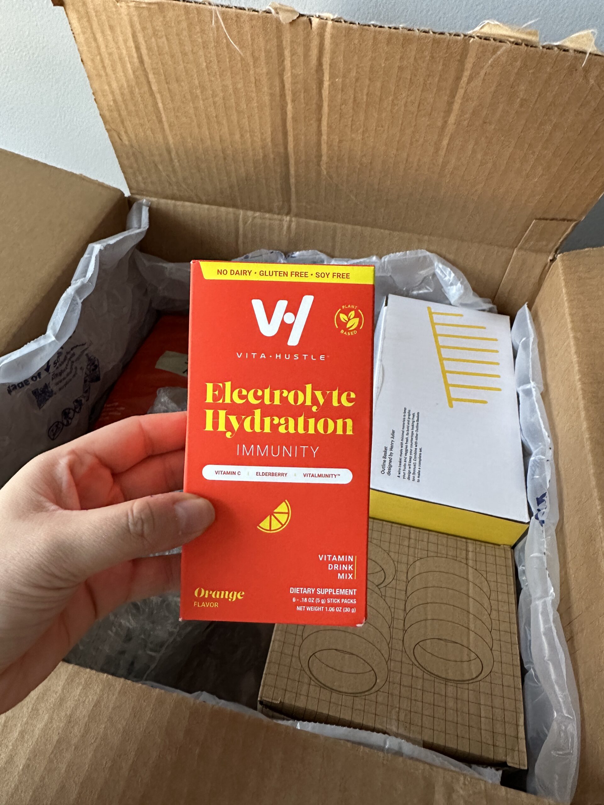 A hand holds a box of VitaHustle Electrolyte Hydration Immunity supplement in orange flavor over an open shipping box containing more products.