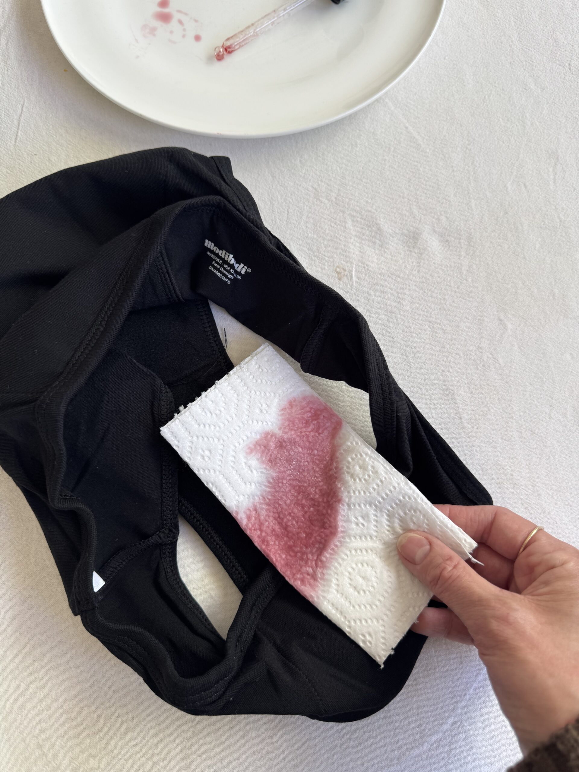 Hand holding a paper towel with a reddish stain next to a black garment on a white surface.
