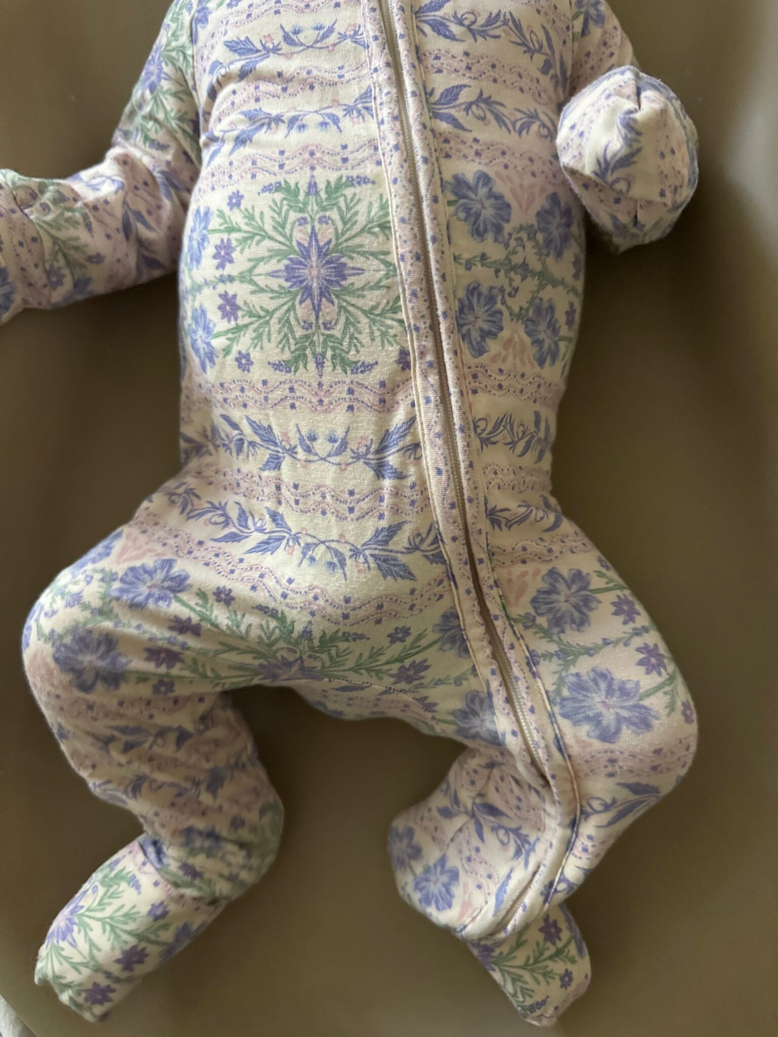 A baby wearing a floral-patterned onesie with a zipper lies on their back; the baby's face is not visible in the image.