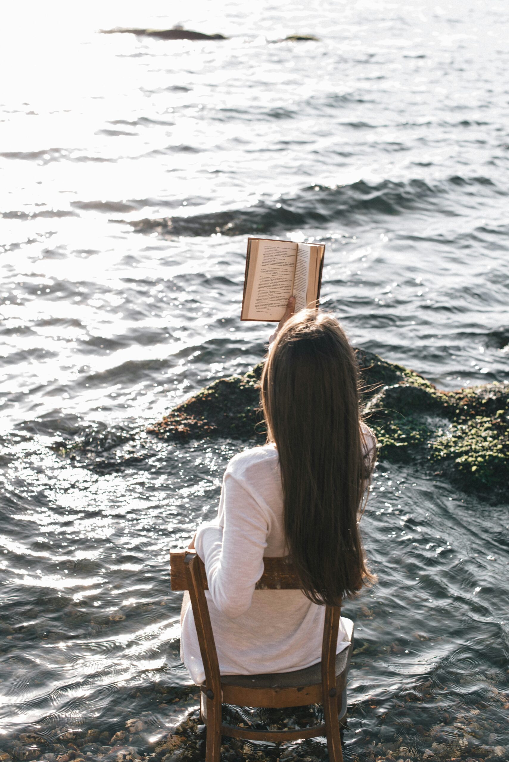 A person with long hair sits on a wooden chair at the edge of the water, reading a book with sunlight reflecting off the sea.