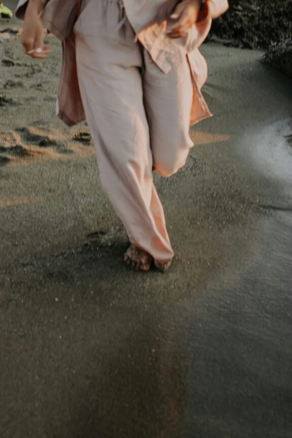 A person wearing loose, light pink clothing walks barefoot along the wet sand at the beach.