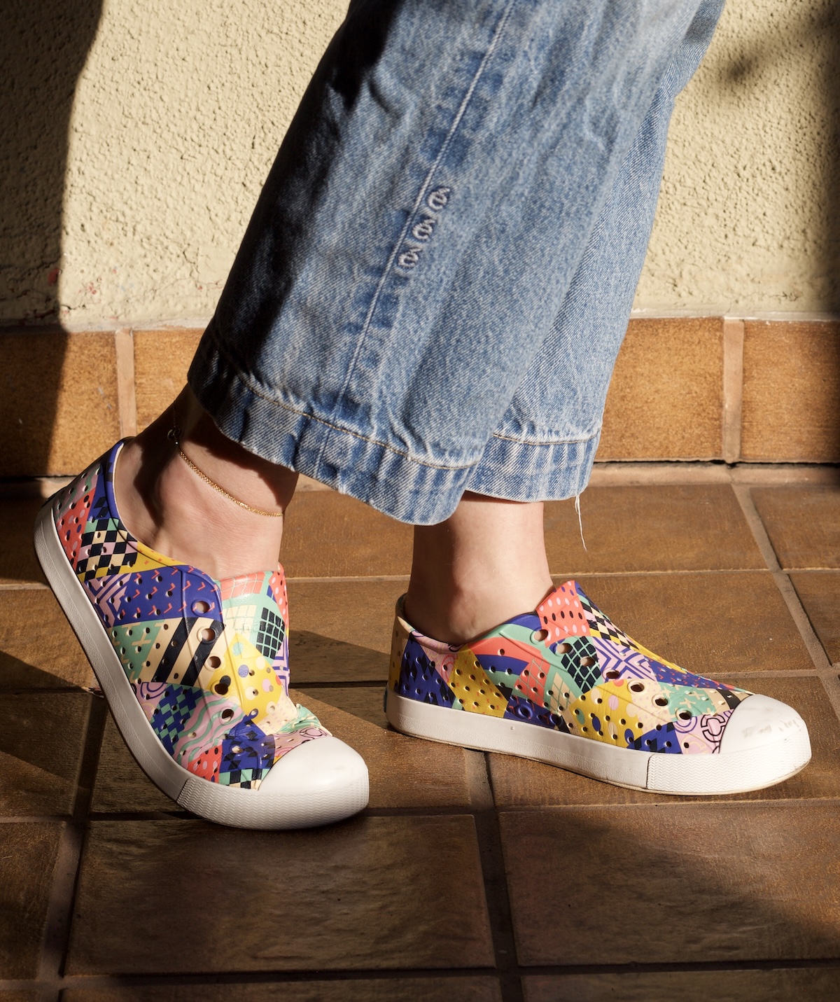 A person wearing colorful sustainable sneakers and blue jeans stands on a tiled floor in sunlight.