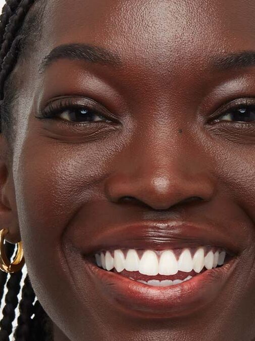 A close-up of a woman's face showing "before" (left) and "after" (right) images highlighting a difference in skin tone applied to the same woman with braided hair and gold hoop earrings.