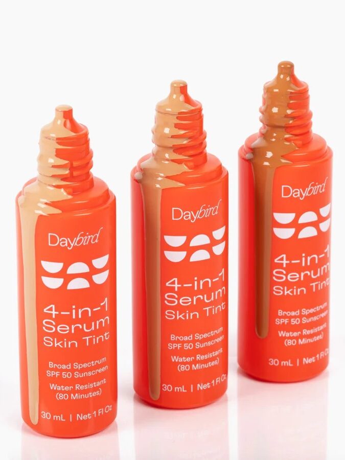 Three orange bottles of Daybird 4-in-1 Serum Skin Tint SPF 50 sunscreen, with shades of tint visible at the tube openings.