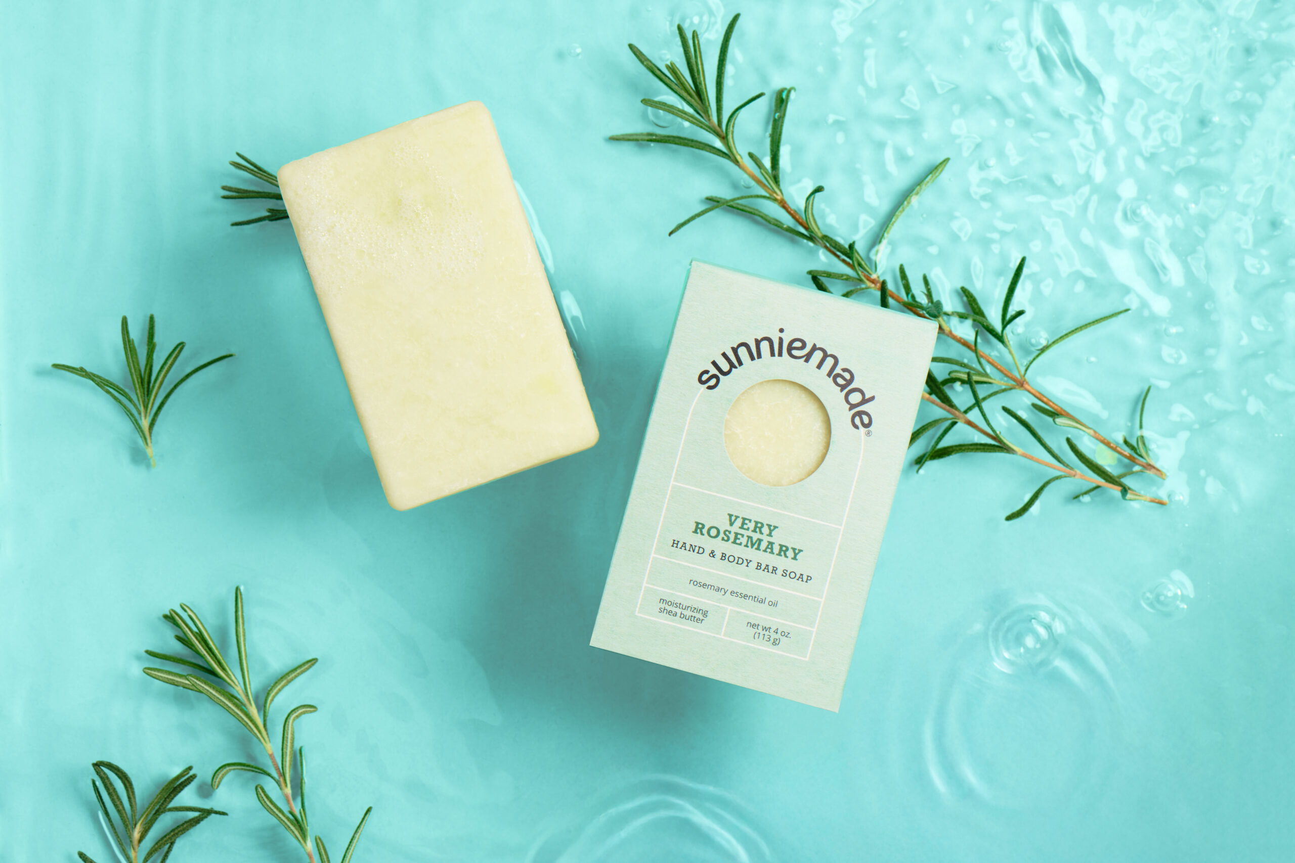 Bar of soap and box labeled "Sunniemade Very Rosemary" on a light blue surface with water and rosemary sprigs.