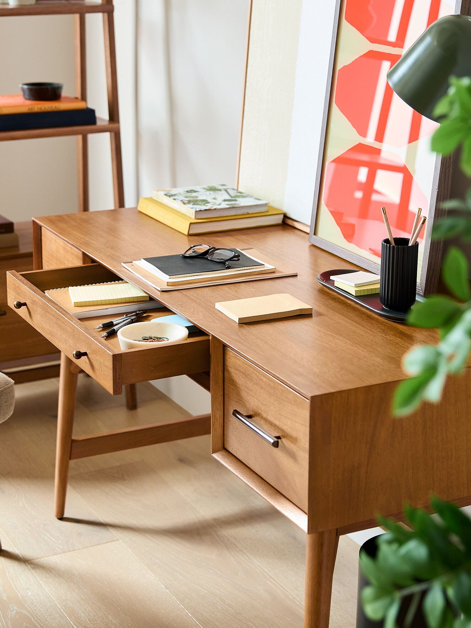 Wooden desk with open drawer holding CDs and stationery, surrounded by books, a lamp, and a framed abstract artwork, next to a beige chair and leafy plant.