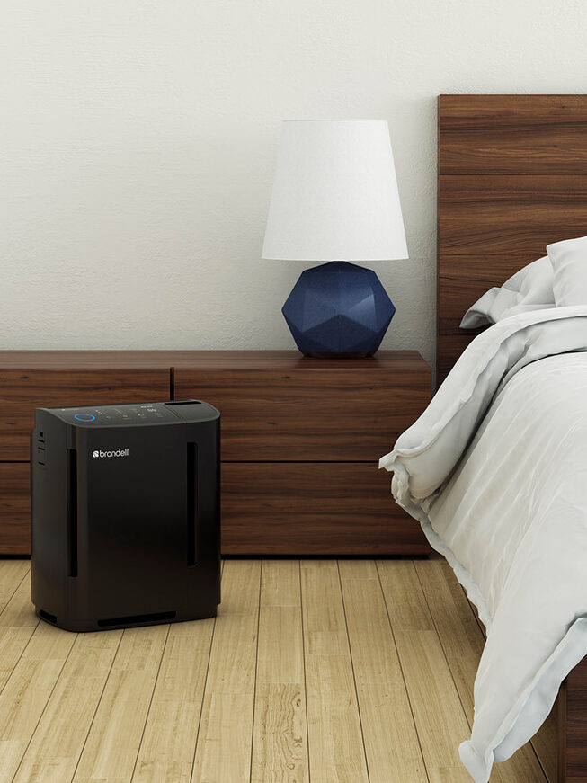 A bedroom with a wooden bed, white bedding, and a nightstand. An air purifier is on the floor next to the nightstand. A blue, geometric lamp sits on the nightstand.