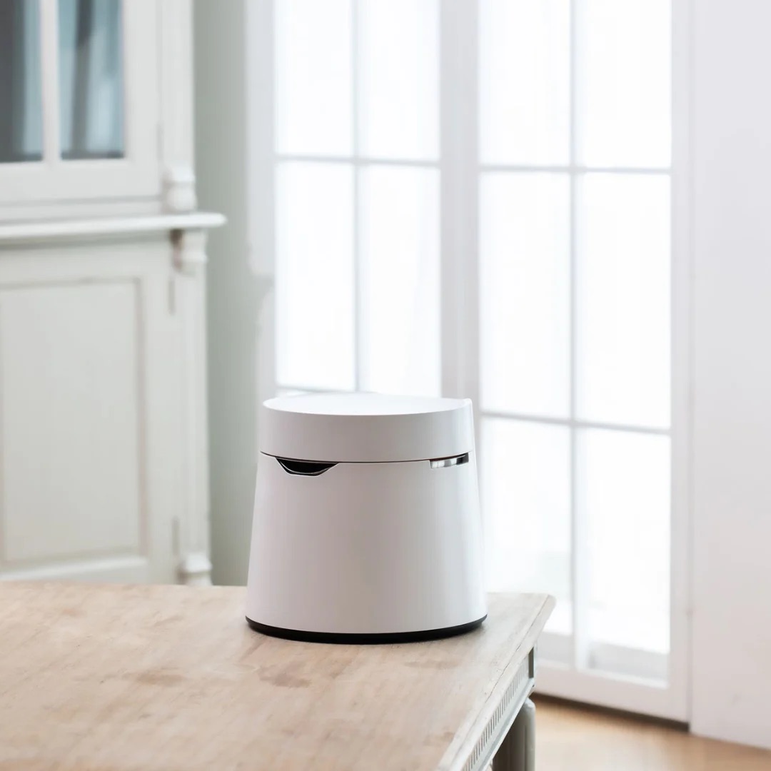 A small, white cylindrical device sits on a wooden table in a bright room with large windows in the background.