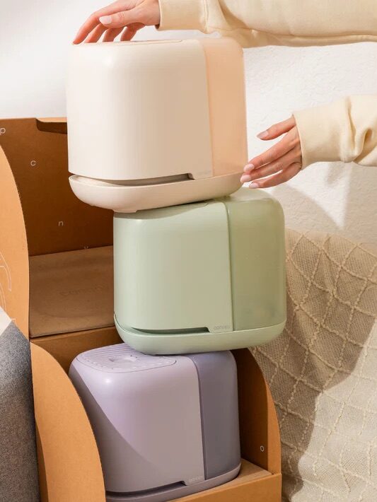 A person places a cream-colored container on top of a stack of pastel-colored containers beside a cardboard box near a crib and chair.