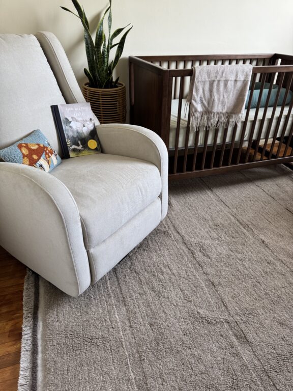Beige armchair with a pillow and book beside a wooden crib, a plant in the background, and a beige rug covering the floor.