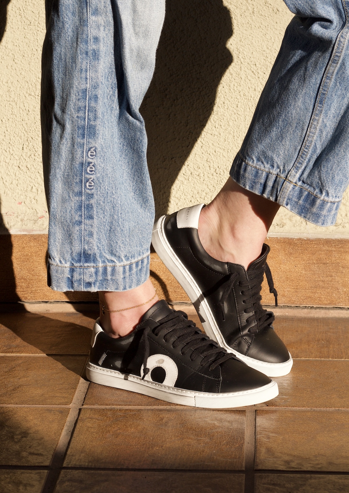 Person wearing rolled-up blue jeans and black sustainable sneakers with a white design, standing on brown tiled floor in sunlight.