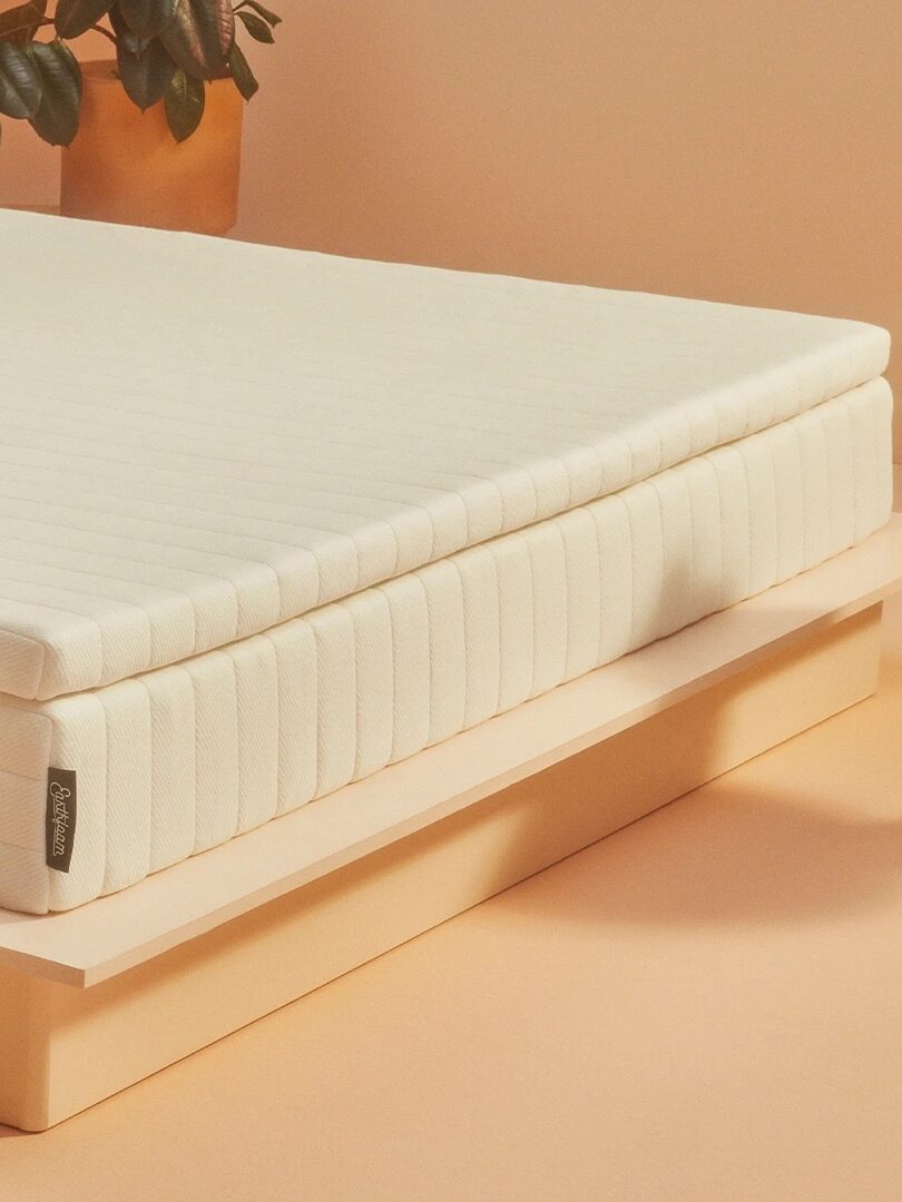 A minimalist bedroom setup featuring a thick, white mattress on a wooden platform bed. There is a round wooden side table with stones on it and a potted plant in the background.