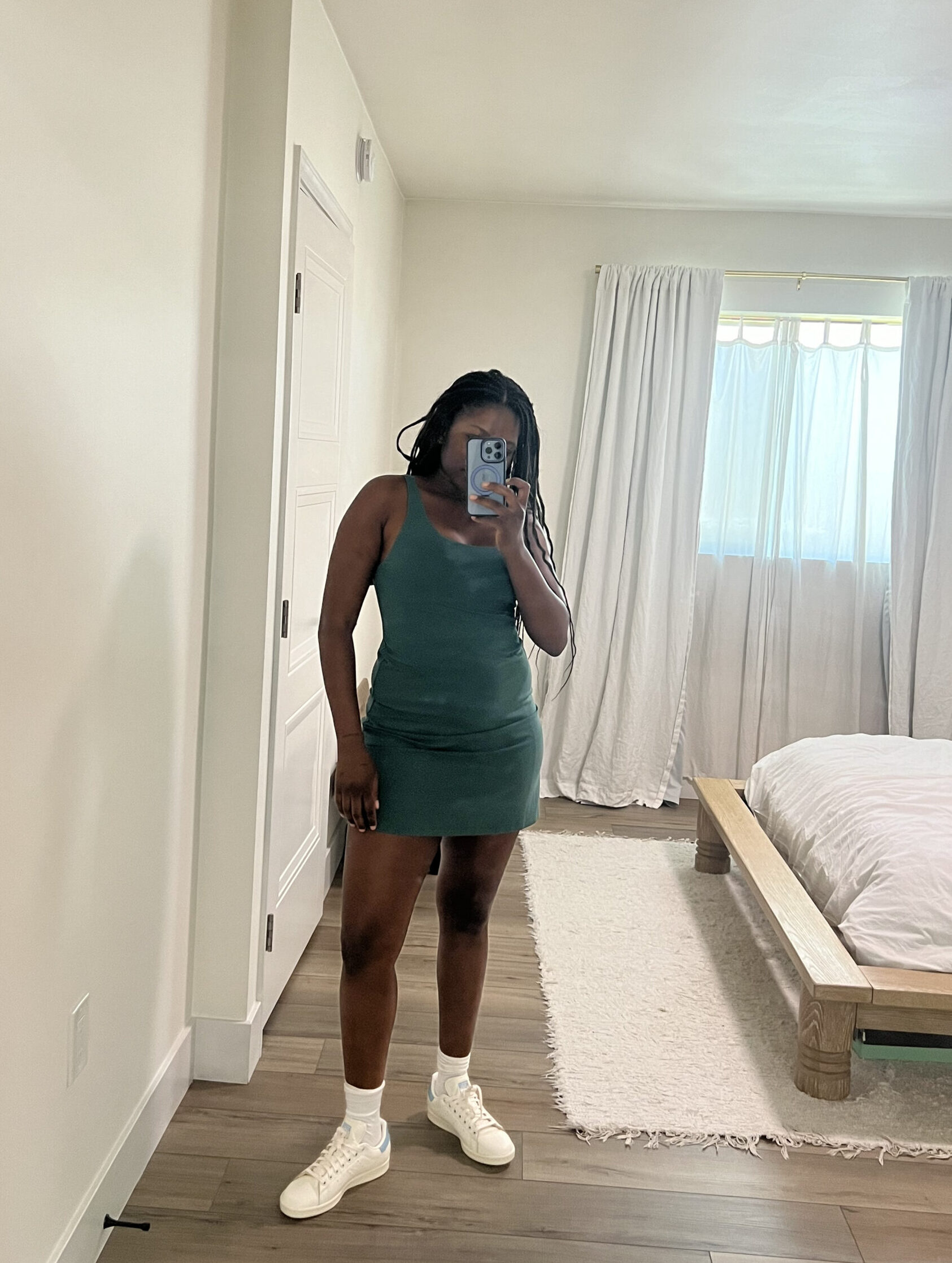 A person in a green dress takes a mirror selfie in a minimalist bedroom. The person is wearing white sneakers. The room has a wooden bed, white curtains, and a white rug.
