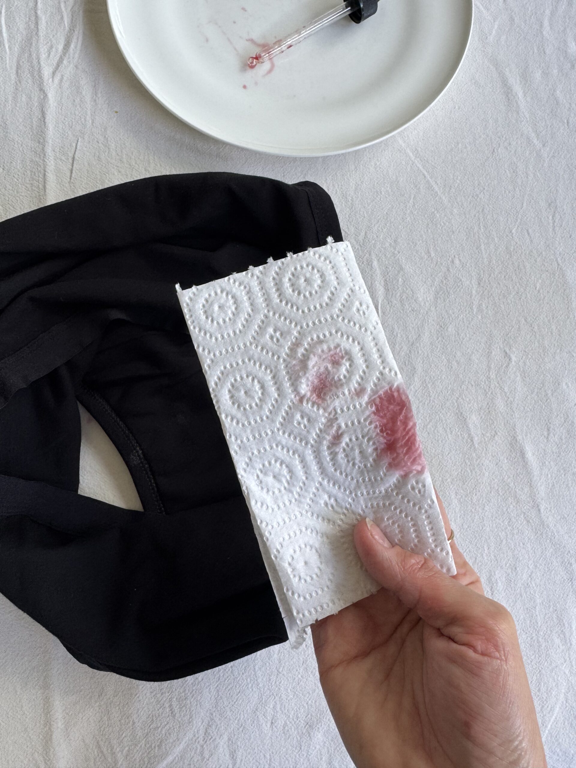 A hand holds a paper towel with lipstick marks. A black garment and a white plate with a dropper and more lipstick stains are visible in the background.