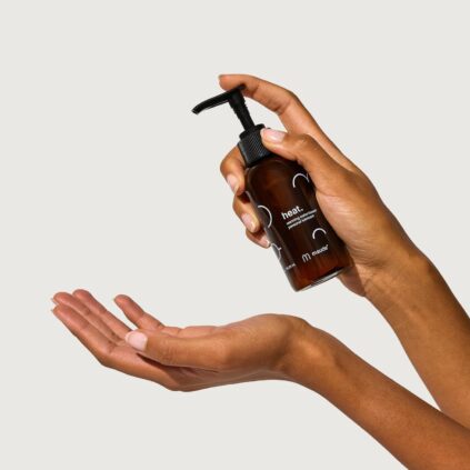 Two hands holding a brown bottle with a pump dispenser, labeled "heat." and "m." One hand holds the bottle upright while the other is positioned to receive the product.