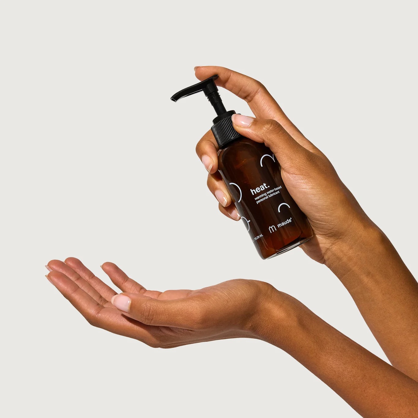 Two hands holding a brown bottle with a pump dispenser, labeled "heat." and "m." One hand holds the bottle upright while the other is positioned to receive the product.