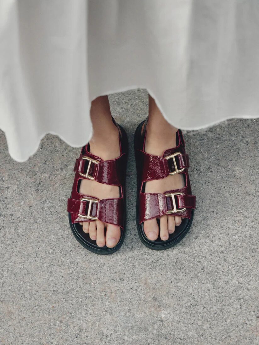 A person wearing a white skirt and burgundy sandals with double buckles stands on a gray concrete surface.