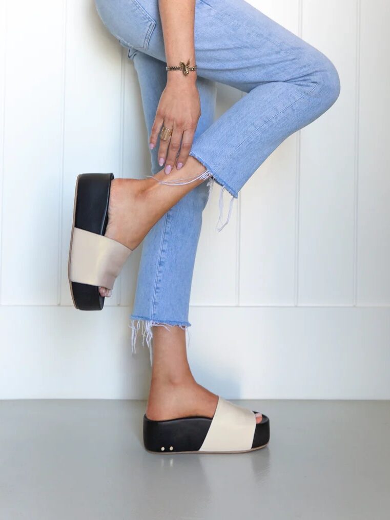 Person wearing frayed light blue jeans and black-and-beige slide sandals stands on one leg, holding the ankle of the other leg, against a white paneled wall.