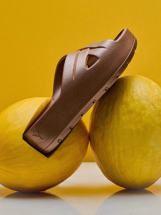 A brown sandal is balanced on top of two yellow melons against a solid yellow background.