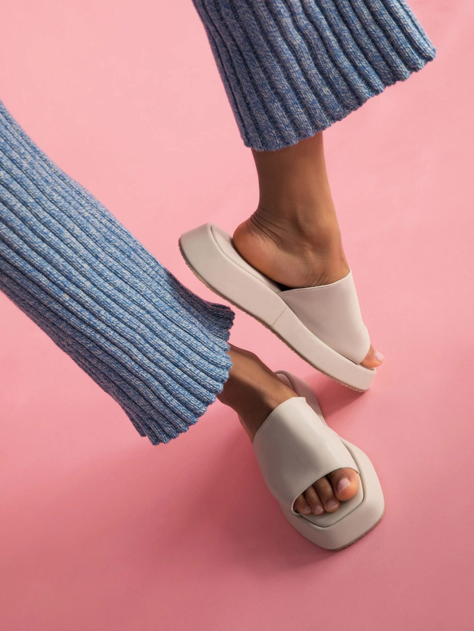 A person wearing ribbed blue pants and beige slide sandals stands against a pink background.