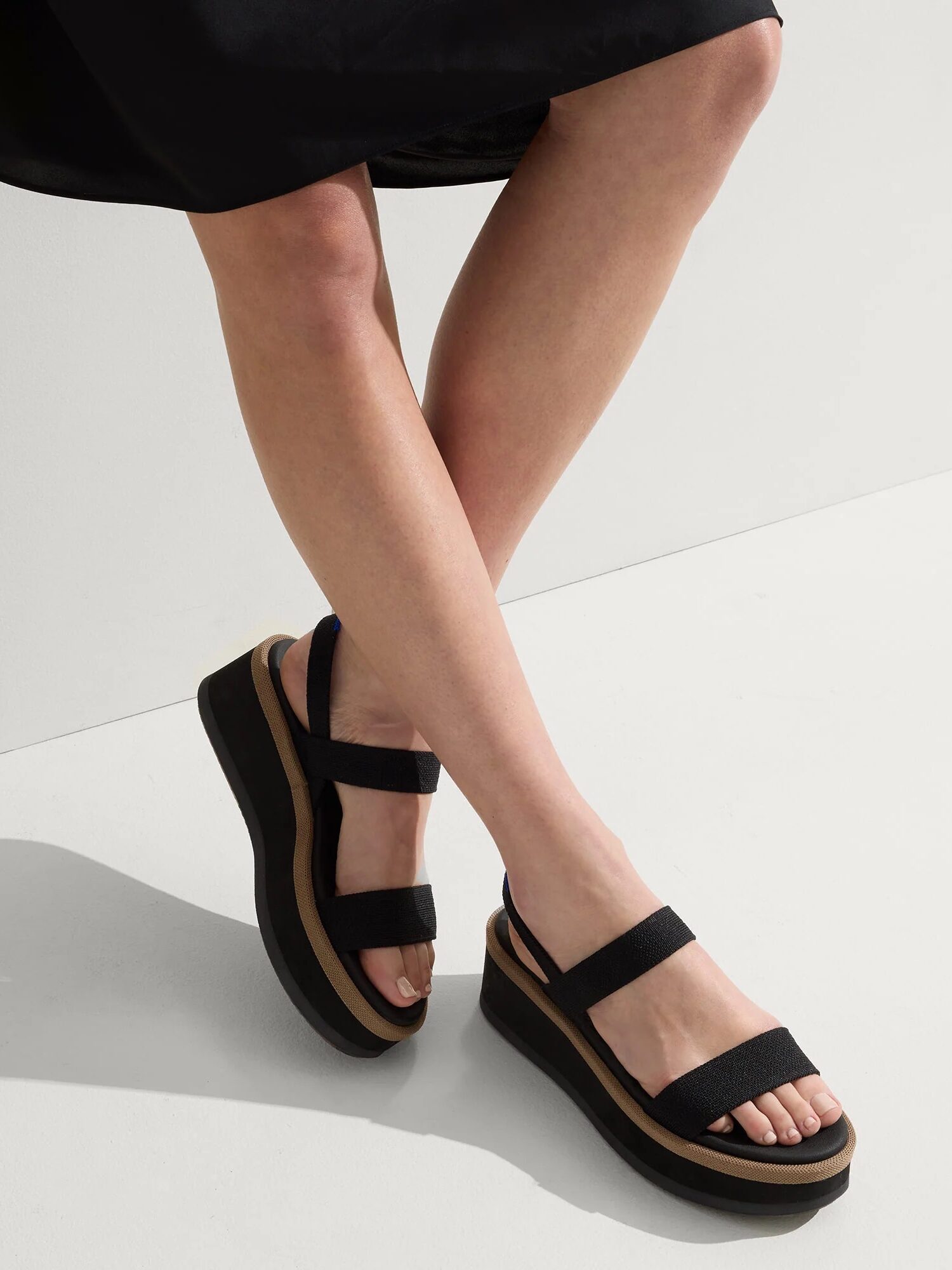 A person wearing a black dress and black platform sandals with two straps stands on a light-colored floor, legs crossed at the ankles.