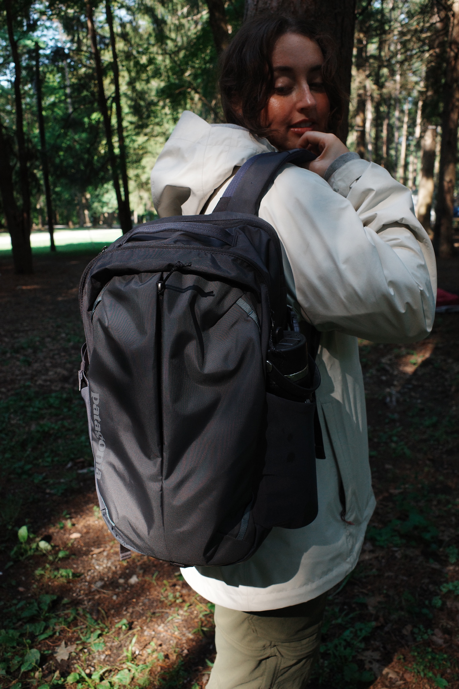 Person wearing a light-colored jacket and carrying a large black backpack stands in a wooded area with green trees and sunlight filtering through.