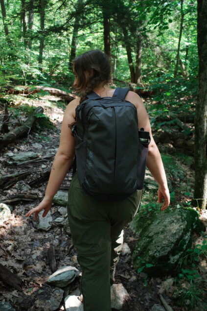 A person wearing a backpack and green pants walks along a rocky, wooded trail surrounded by trees and greenery.