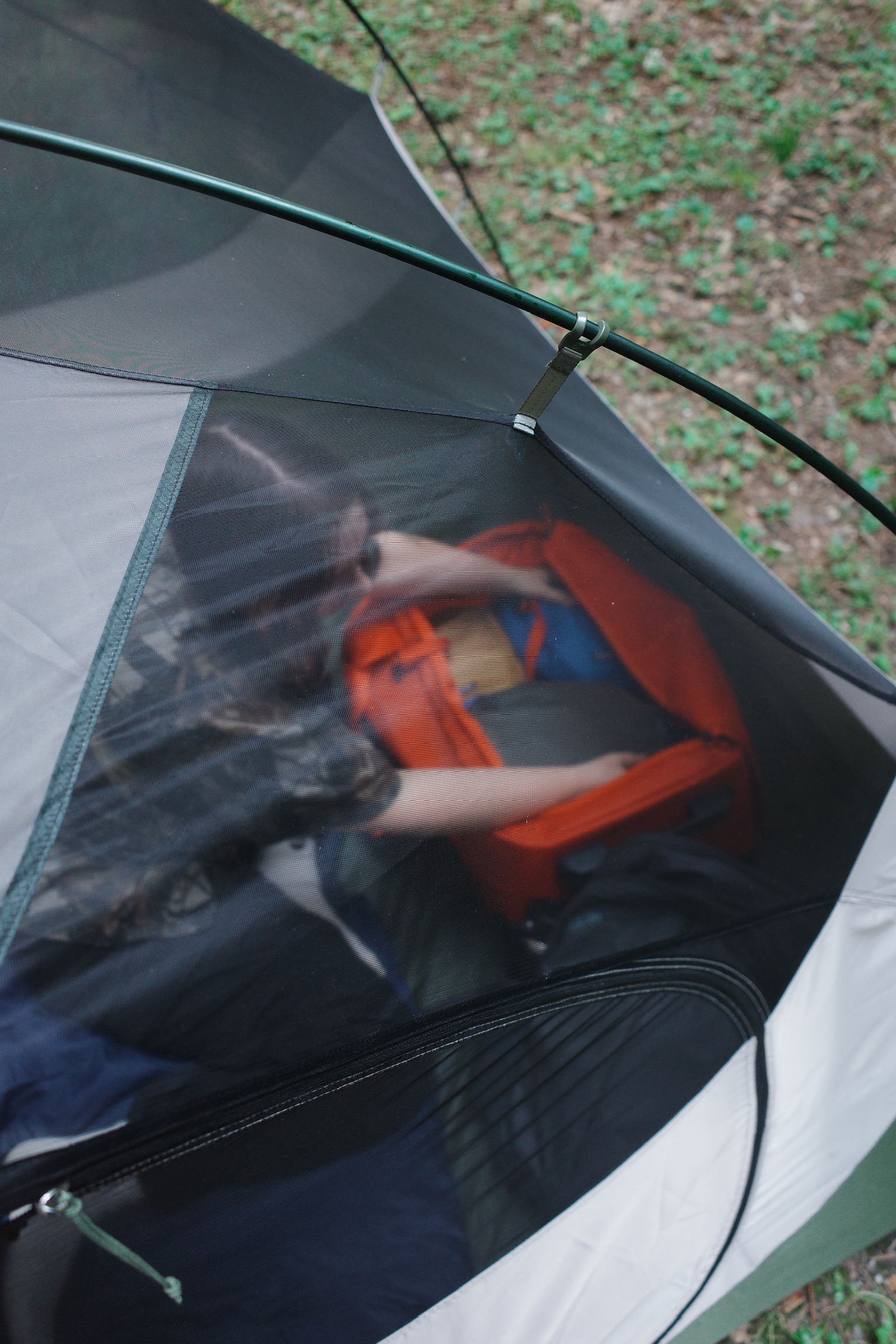 Person seen through a tent mesh organizing supplies inside an orange container, with grass and leaves visible outside the tent.