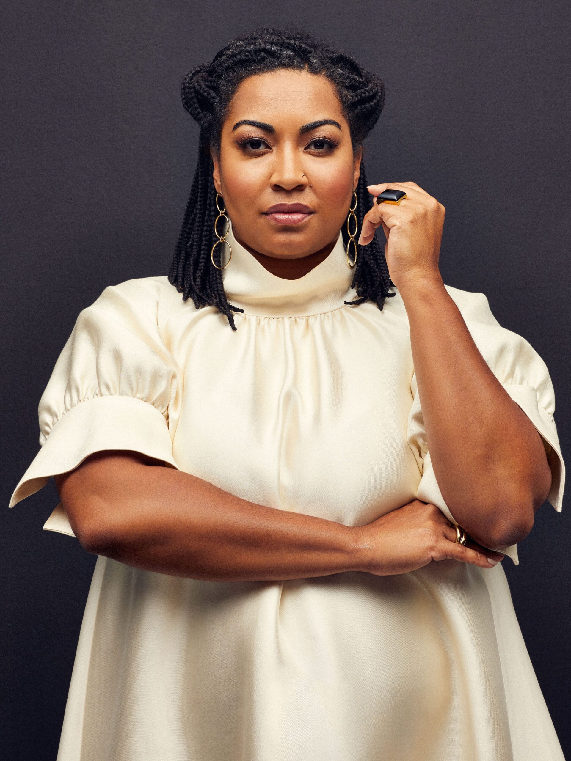 A woman in a cream-colored dress stands against a dark background, looking at the camera with one arm crossed and the other hand touching her hair.