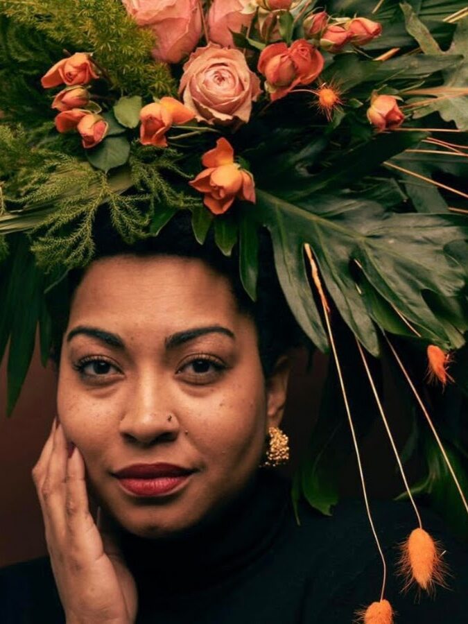 A woman with a calm expression wears a large headdress made of green leaves, orange roses, and various foliage, with her hand resting on her face.