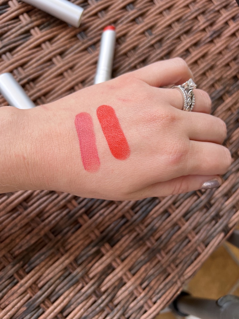 A hand with two lipstick swatches on the back, one pink and one red, displayed over a wicker surface. Several tubes of lipstick are visible in the background.