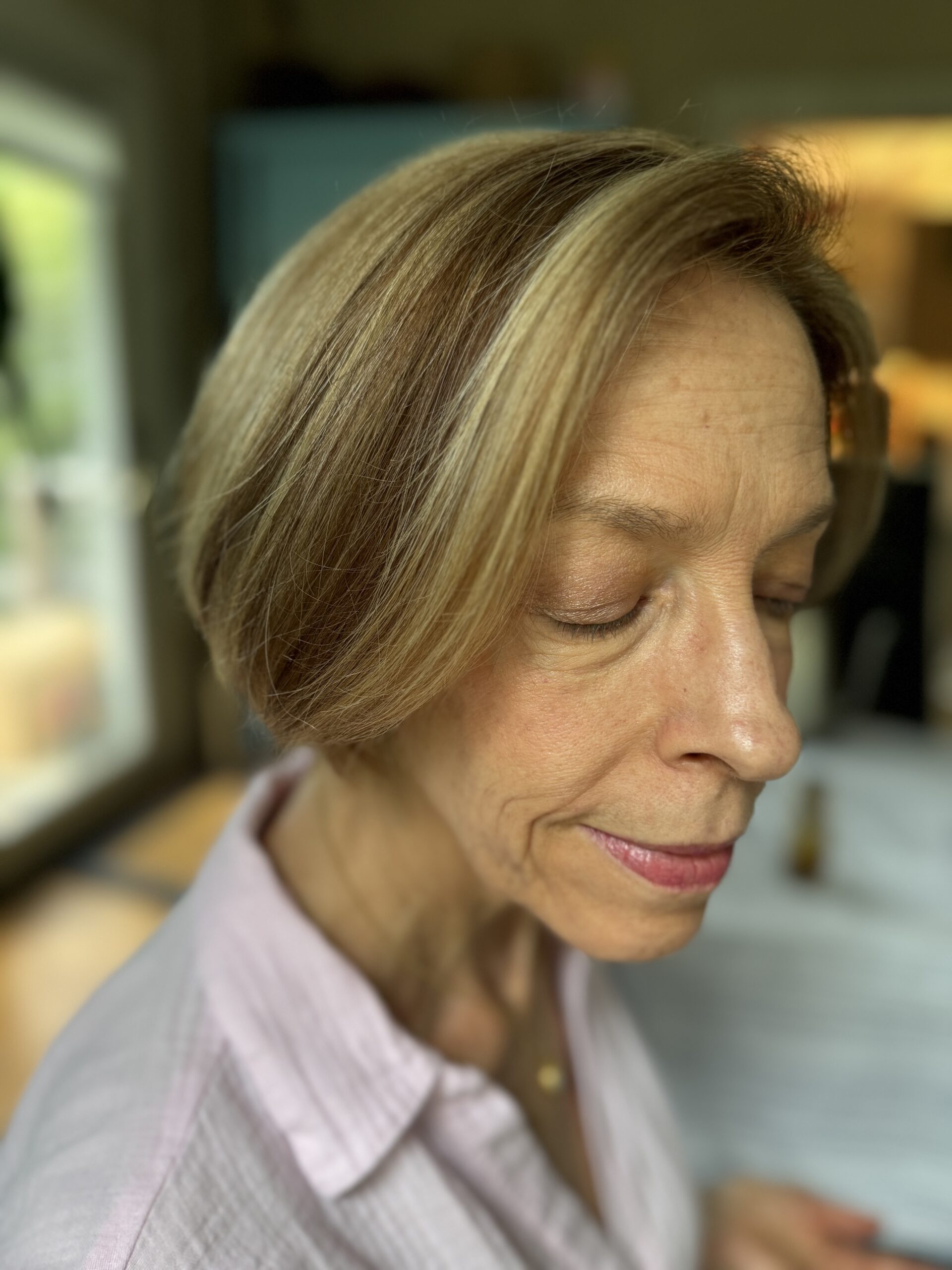 A close-up of an older woman with short, light brown hair, wearing a pink shirt, looking down with her eyes closed. The background is slightly blurred.