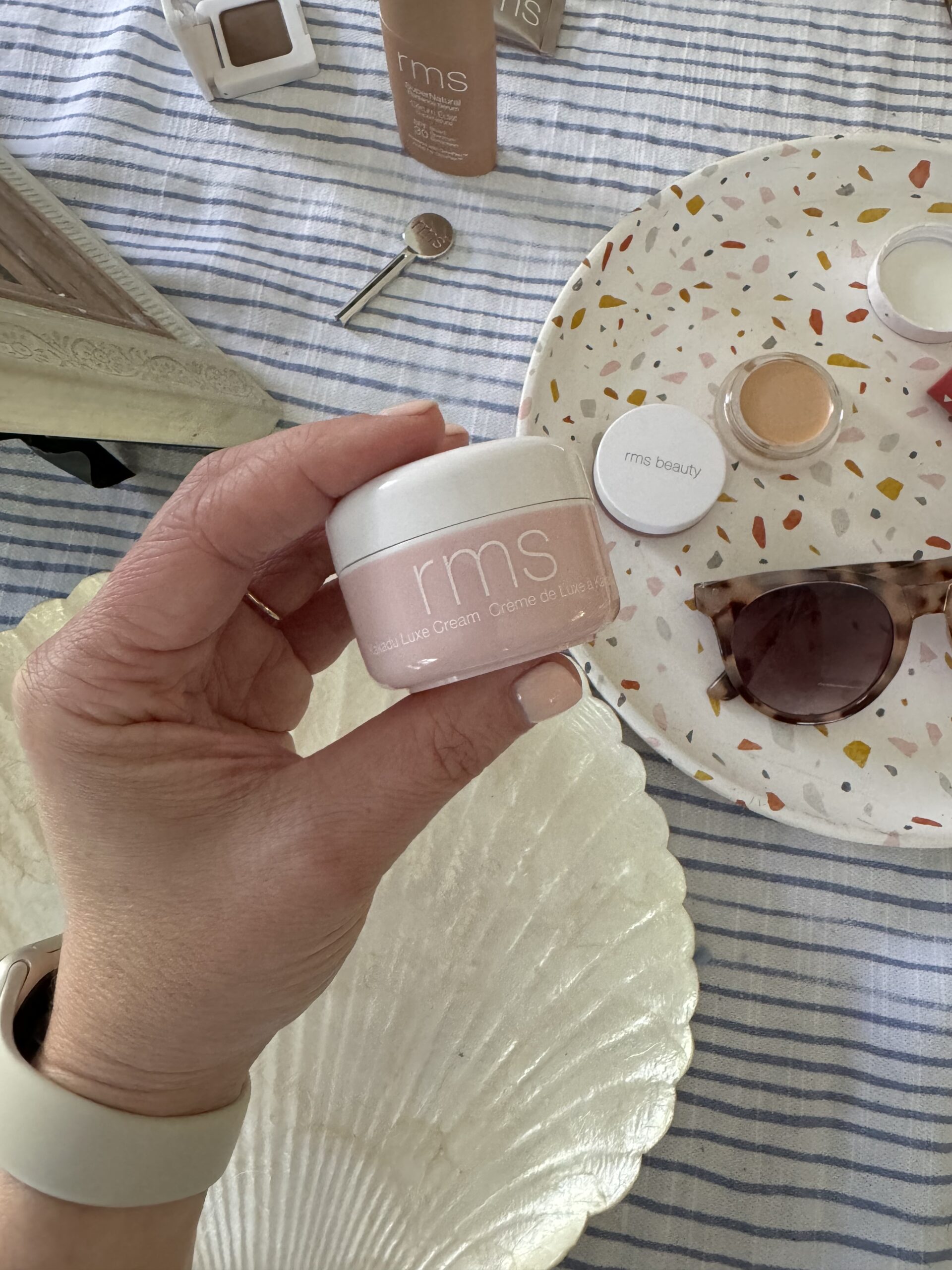A hand holds a jar of "rms" beauty face cream above a table with a compact mirror, makeup items, sunglasses, and a shell-shaped dish.